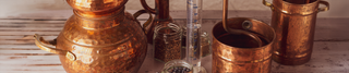 A traditional copper distillation setup with jars of juniper berries and other botanicals, a glass measuring cylinder, and copper components, set on a rustic wooden surface