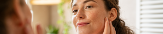 A young woman with curly brown hair and a radiant complexion gently touches her face while admiring her reflection in the mirror. She stands in a softly lit bathroom with warm, natural tones, surrounded by potted plants and wooden shelves. Her serene expression conveys confidence and self-care.