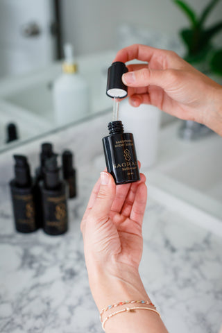 Hand holding Night Bakuchiol Serum bottle with dropper over marble countertop and skincare bottles in background.