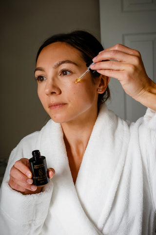 Woman in white robe applying Night Bakuchiol Serum with dropper to her face in a skincare routine.