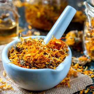 A white ceramic mortar and pestle filled with dried calendula flowers, placed on a rustic burlap surface. Dried petals are scattered around, with glass jars containing more dried herbs visible in the background.