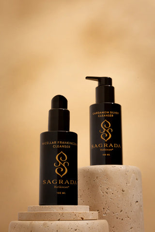 The Dual Cleanser Bundle from Sagrada, featuring Micellar Frankincense Cleanser and Cardamom Silk Cleanser in black bottles with gold branding, elegantly displayed on stone pedestals against a warm beige background