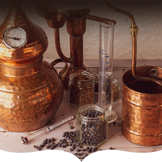 A traditional copper distillation setup with jars of juniper berries and other botanicals, a glass measuring cylinder, and copper components, set on a rustic wooden surface