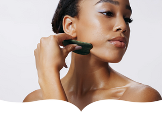 A woman with radiant skin using a jade gua sha stone to massage her face, demonstrating a self-care skincare routine against a clean white background.