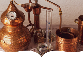 A traditional copper distillation setup with jars of juniper berries and other botanicals, a glass measuring cylinder, and copper components, set on a rustic wooden surface