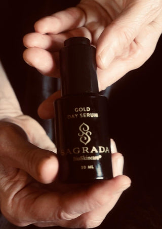 Hands holding a black bottle of Sagrada Gold Day Serum, featuring elegant gold lettering, against a dark background.