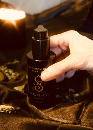 A hand holding a black bottle of Sagrada Cardamom Silk Cleanser, set against a dark, luxurious backdrop with a lit candle and natural stones.