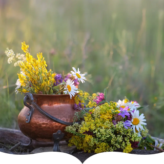 A copper pot filled with freshly picked wildflowers, including daisies and yellow blooms, set against a lush green meadow background.