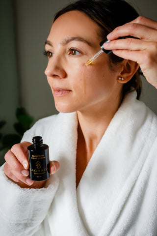 Woman in white robe applying serum to face from bottle, showcasing Award Winning Essentials skincare product.