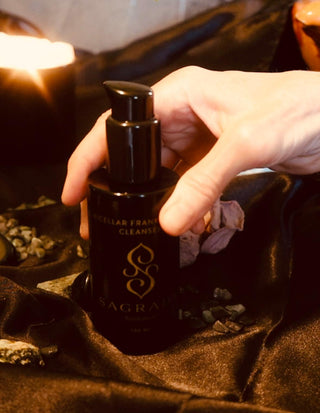 A hand holding a black bottle of Sagrada Micellar Frankincense Cleanser, set on a luxurious dark fabric background with a lit candle and natural stones.