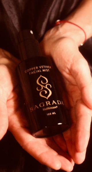 Hands gently holding a black bottle of Sagrada BioSkincare  Copper Vetiver Facial Mist, featuring gold branding and set against a dark, elegant background.