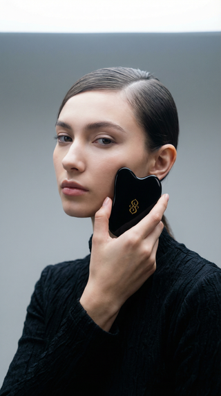 Model using Sagrada black gua sha tool on cheek for facial massage, promoting lymphatic drainage and skin sculpting