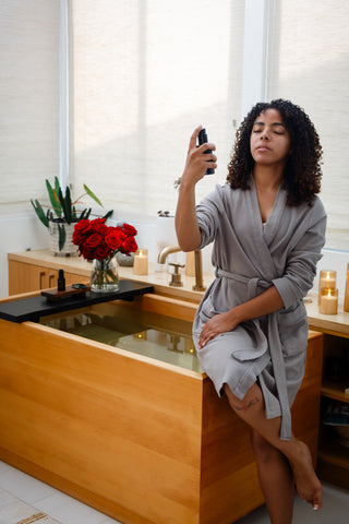 Woman in a bathrobe misting her face with Sagrada facial mist in a candlelit bathroom, creating a calming and luxurious self-care ritual