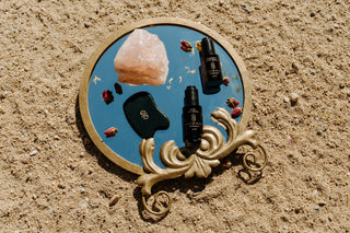 Sagrada BioSkincare Gold Day Serum and Bakuchiol Night Serum styled with a rose quartz crystal, gua sha, and dried botanicals on a decorative mirror in the sand.