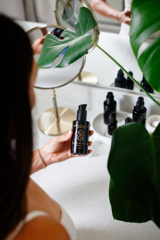 Woman holding Sagrada Micellar Frankincense Cleanser at her vanity, surrounded by skincare essentials in a modern, plant-filled bathroom setting