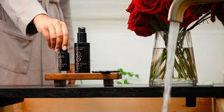 Luxury bath setup featuring Sagrada BioSkincare products, red roses, candles, and a wooden soaking tub in a serene, light-filled bathroom.