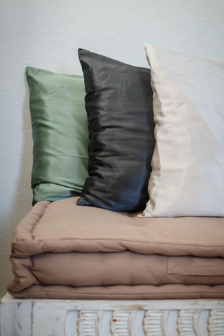 Set of silk pillowcases in sage green, charcoal, and ivory, styled on a folded beige blanket, creating a calming and natural home decor palette.