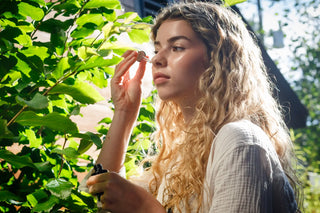 Young woman with glowing skin applying facial serum with a dropper outdoors, surrounded by lush greenery and sunlight, embodying a natural skincare ritual.