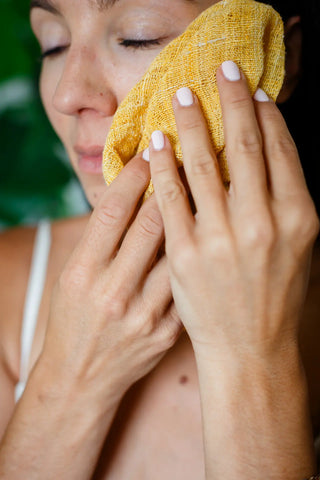 Woman gently cleansing her face with a yellow silk washcloth, eyes closed in a calm moment of her natural skincare ritual.